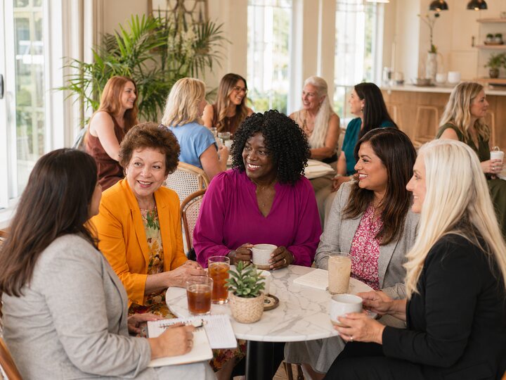 A diverse group of women networking together around a table in a bright casual setting.